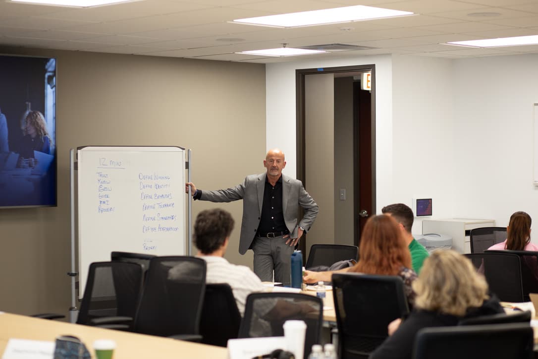 Steve Lowisz in a gray jacket teaching at a whiteboard with participants seated around tables